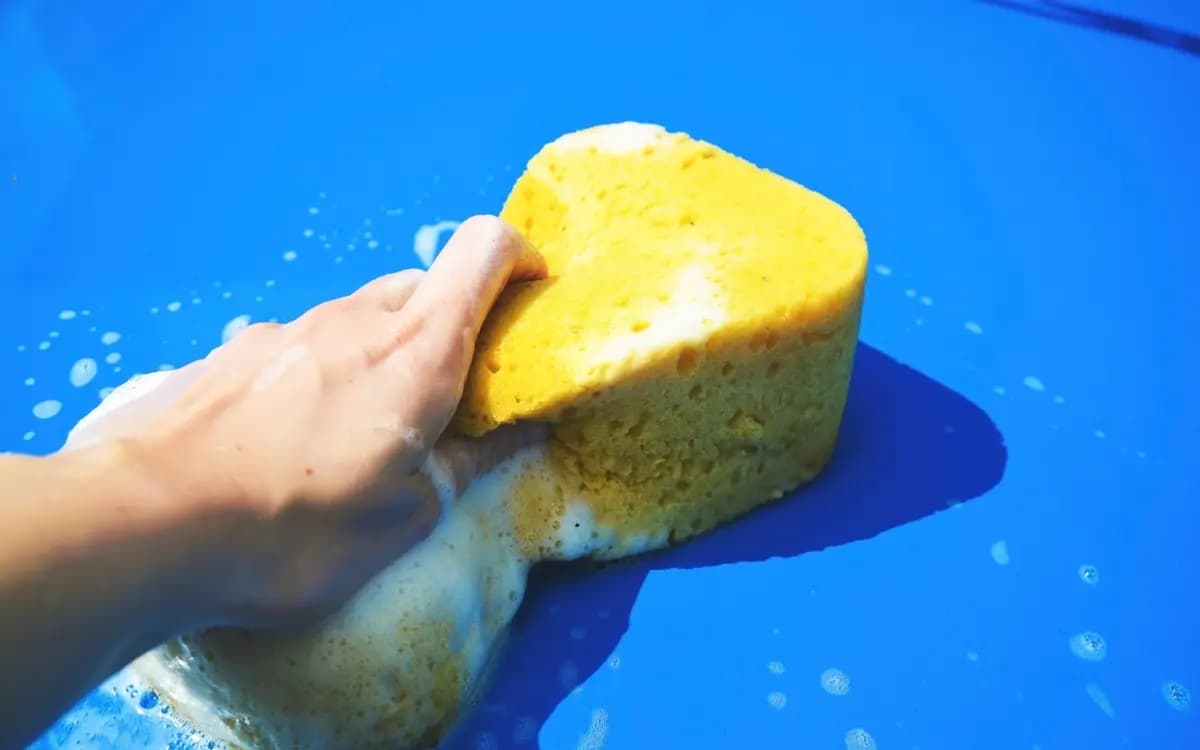 Hand washing a bright blue car with a yellow sponge and white foam